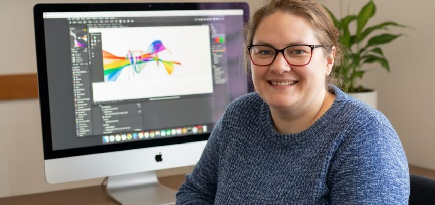 An AI generated photograph of me sitting at a desk. I'm dress casually in a blue jumper and behind me is a screen showing an illustration.